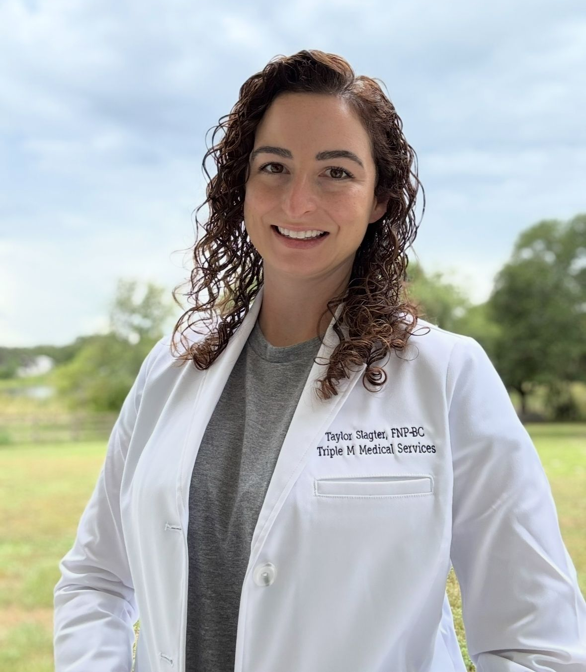 A medical professional with curly hair wearing a white lab coat and gray shirt, smiling outdoors against a natural backdrop.