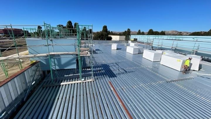Rooftop construction site with corrugated metal decking, scaffolding, and stacked white materials under a blue sky
