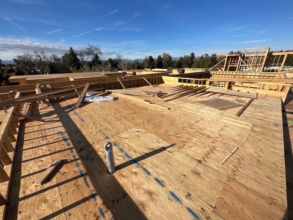 Wooden house framing under construction on a sunny day, with exposed roof trusses and plywood subfloor.