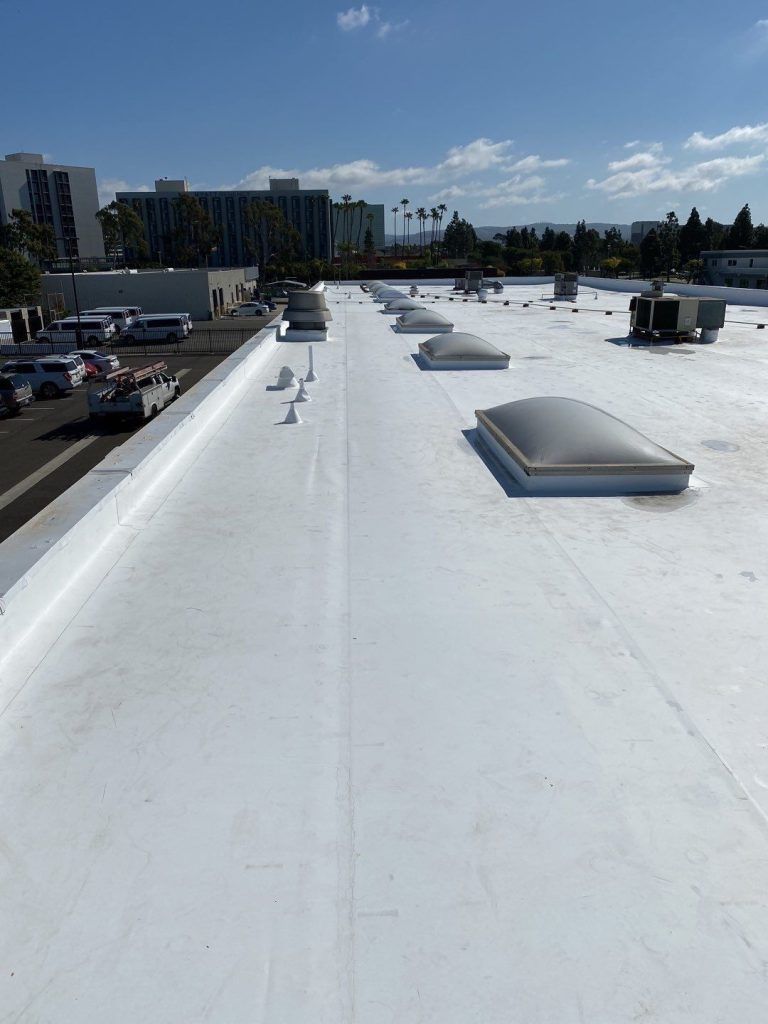 White flat rooftop with skylights, vents, and distant buildings under a clear blue sky.