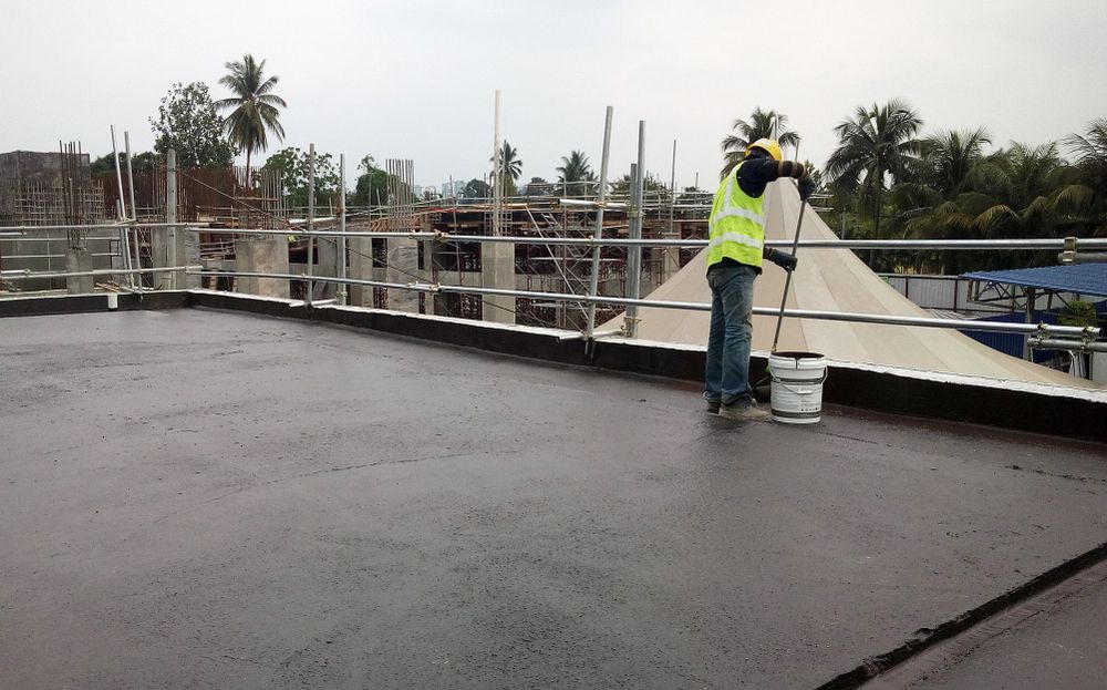Worker in a yellow safety vest on a rooftop, painting or coating the surface near white railings.