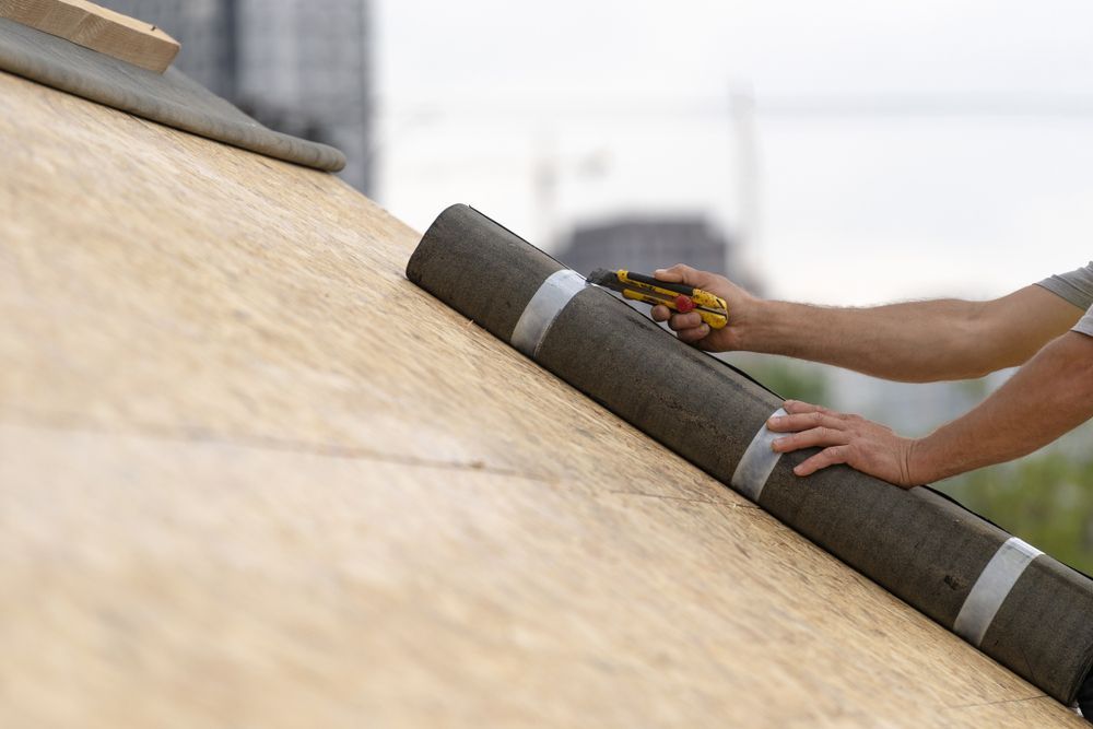 Person laying dark roofing material on a sloped roof with flowers nearby