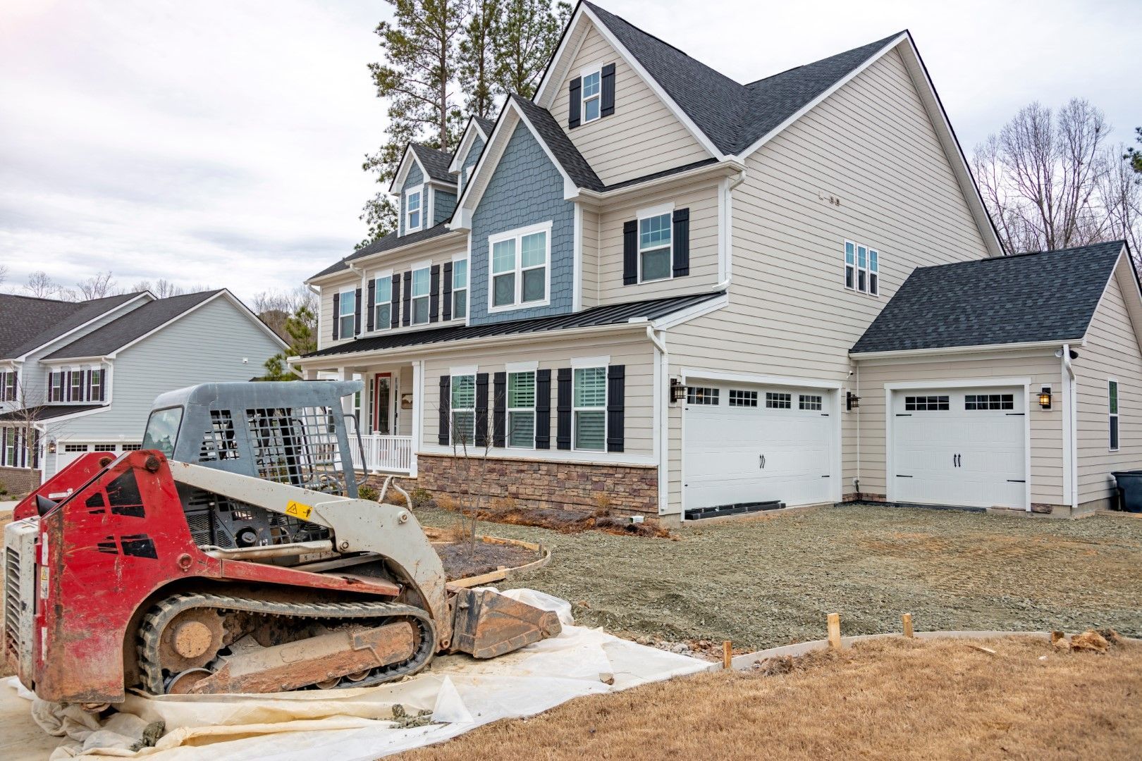 Red and gray skid steer in front of a two-story house under construction with two-car garage and blue siding.