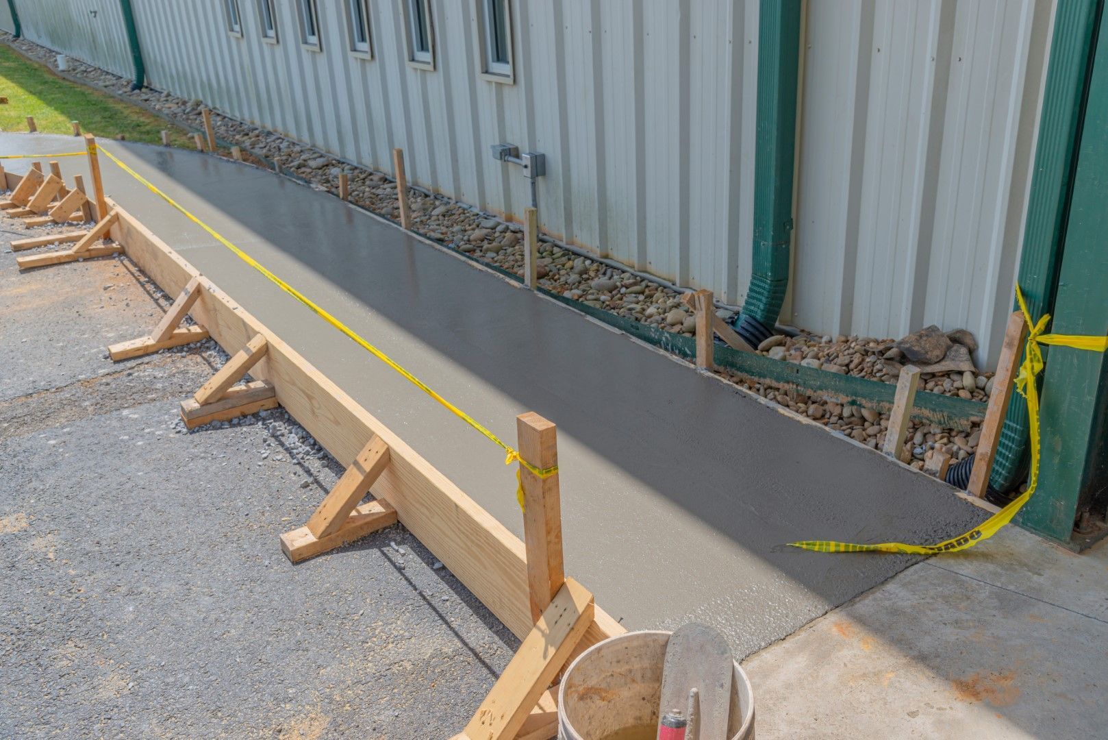 Newly poured concrete walkway next to a building, enclosed by wooden forms and caution tape.