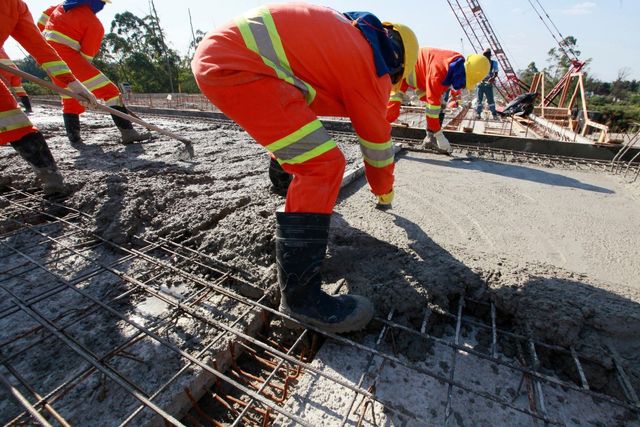 Construction workers in orange coveralls pouring concrete on a rebar framework.