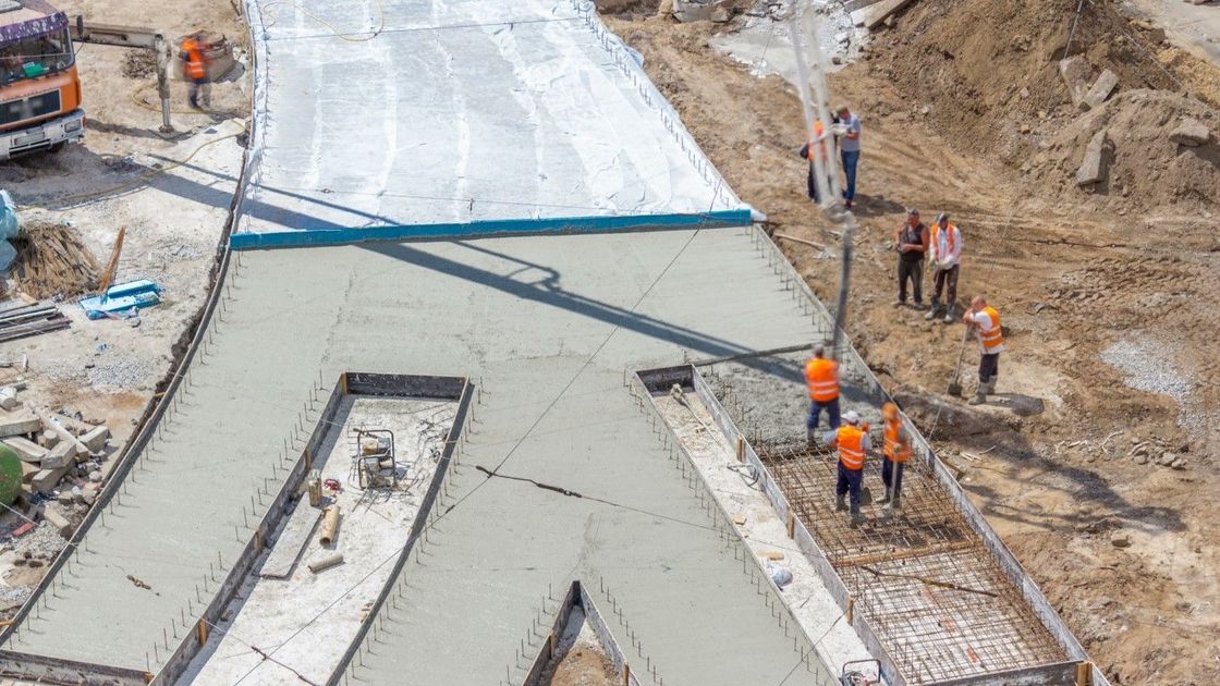 Construction site: workers pouring concrete on a large, abstract concrete structure. Orange vests, dirt, and machinery.