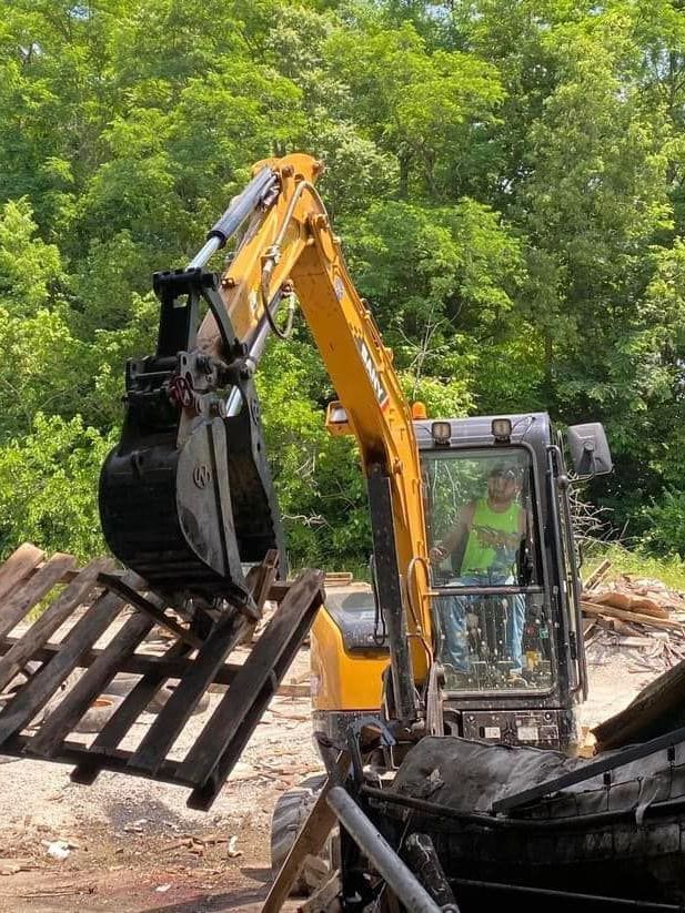A man is driving a yellow excavator carrying a wooden pallet.