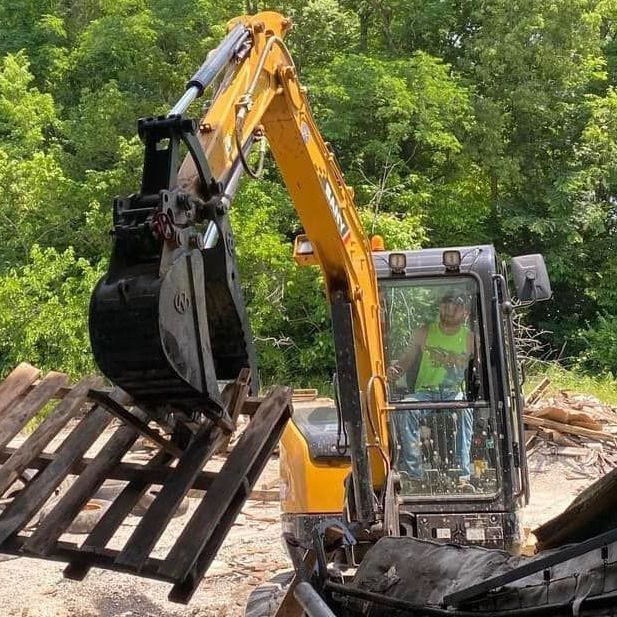 A man is driving a yellow excavator carrying a wooden pallet.