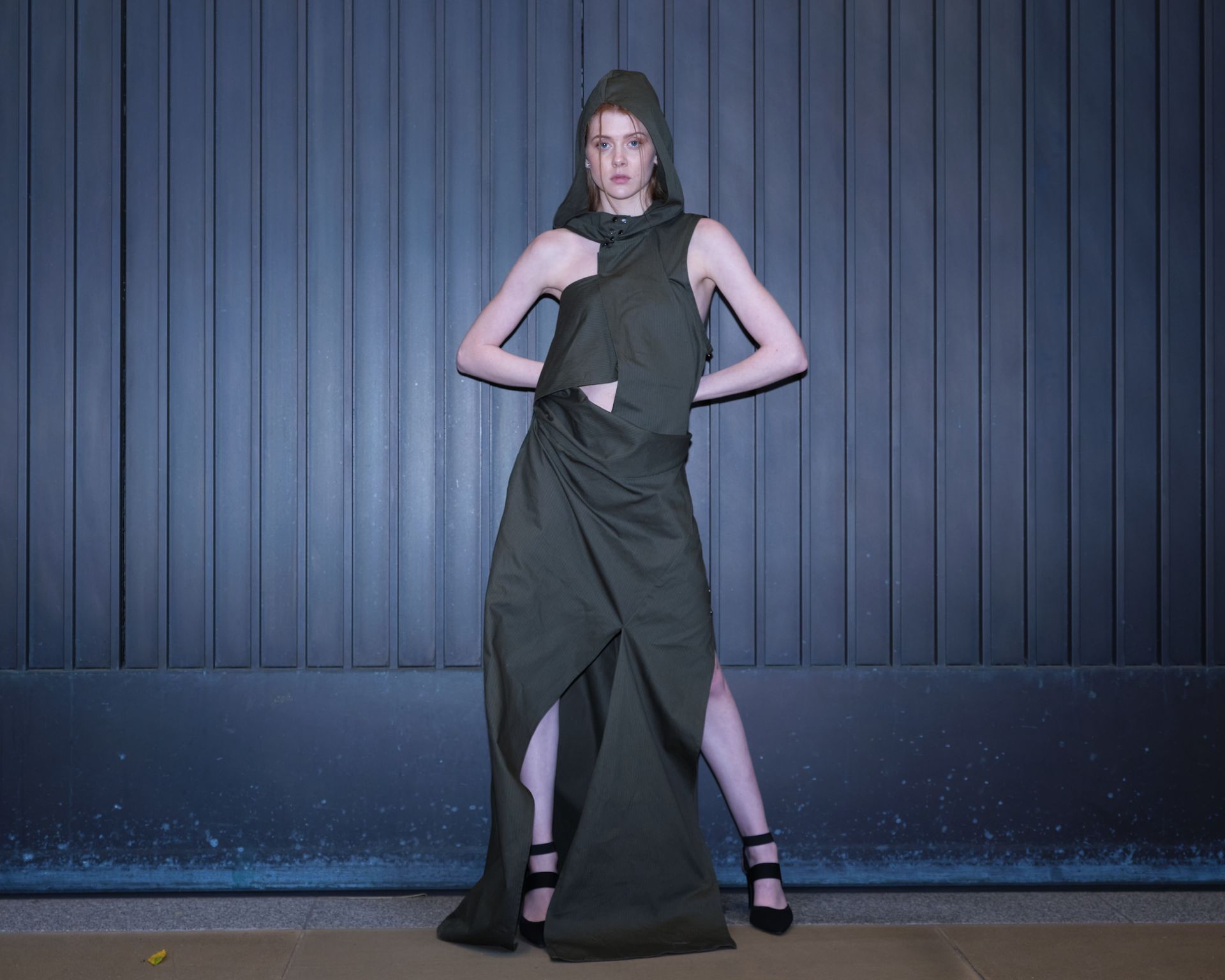 A woman in a long green dress with a hood is standing in front of a wall.