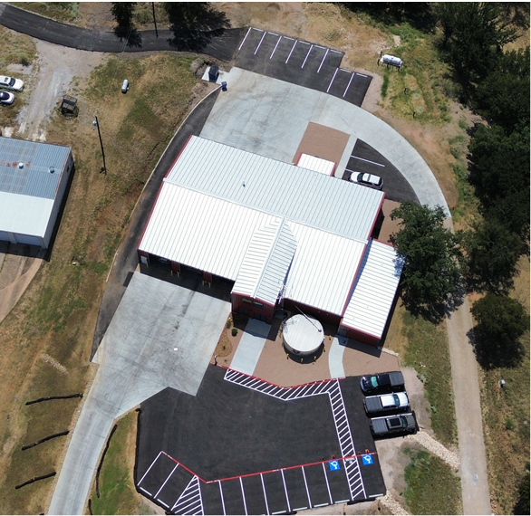 Aerial view of a building with a parking lot, driveway, and surrounding trees.