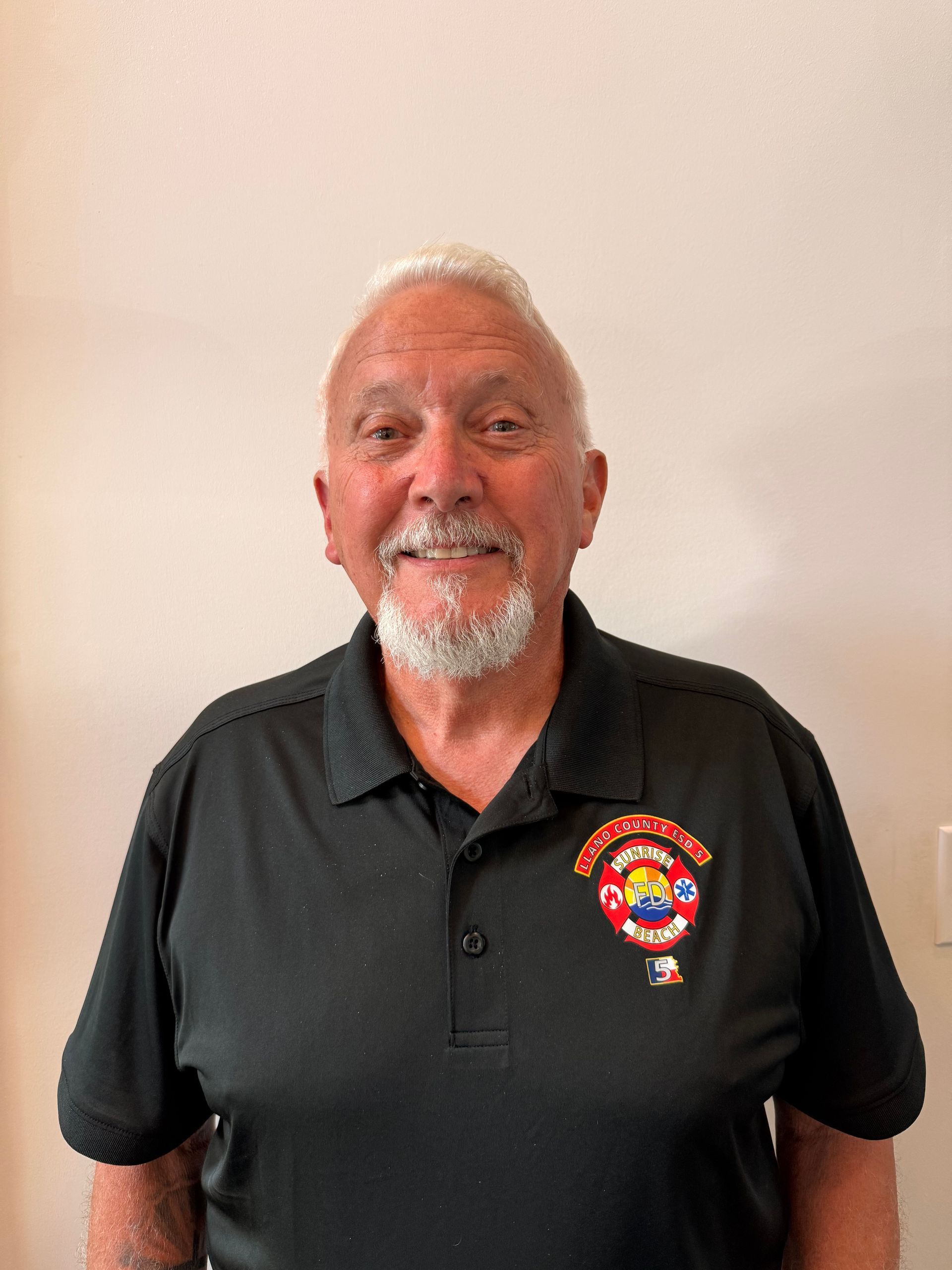 Smiling man in a black polo shirt with a colorful emblem, standing against a plain beige wall.