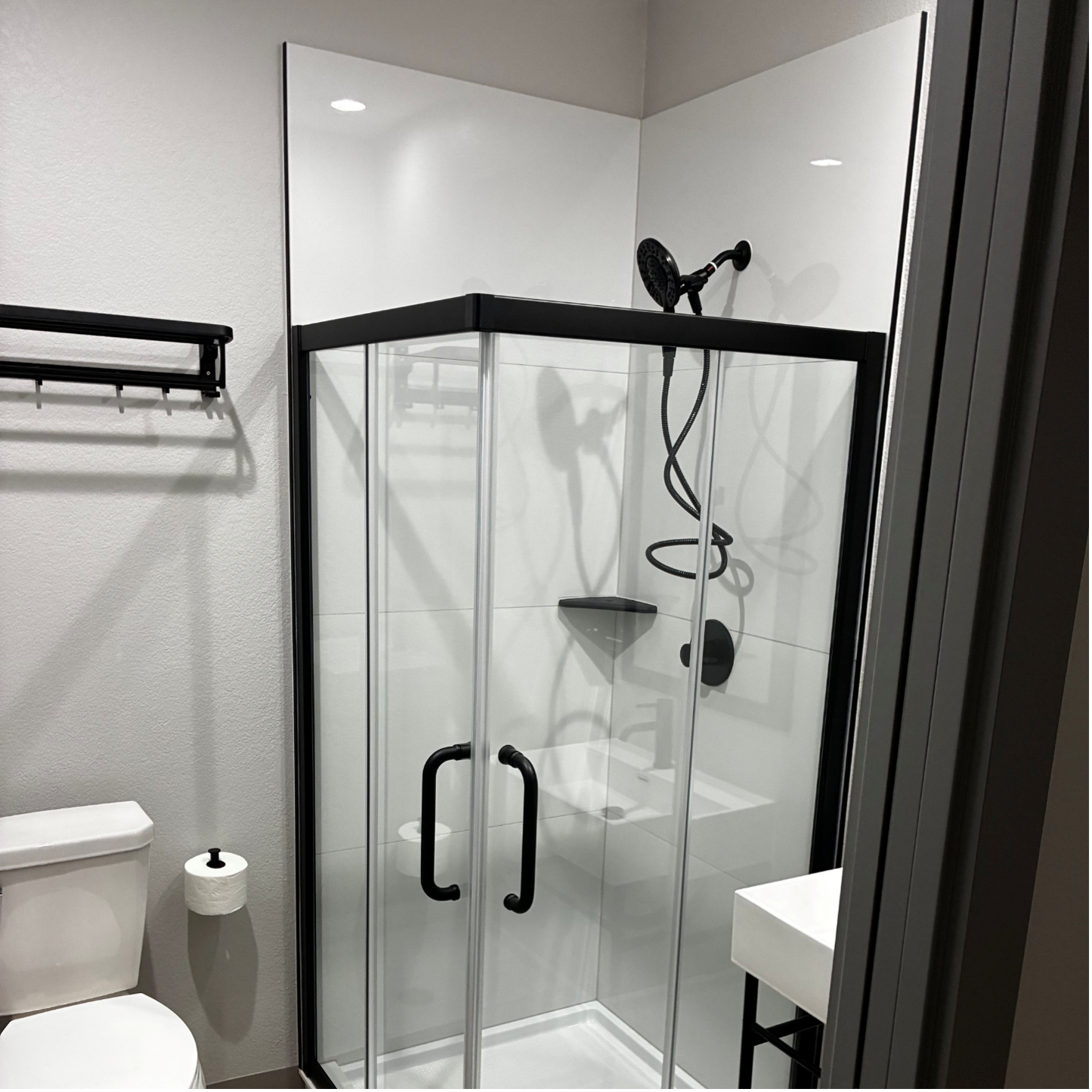 A bathroom with a corner shower featuring a black frame and white walls, next to a toilet and a wall-mounted rack.