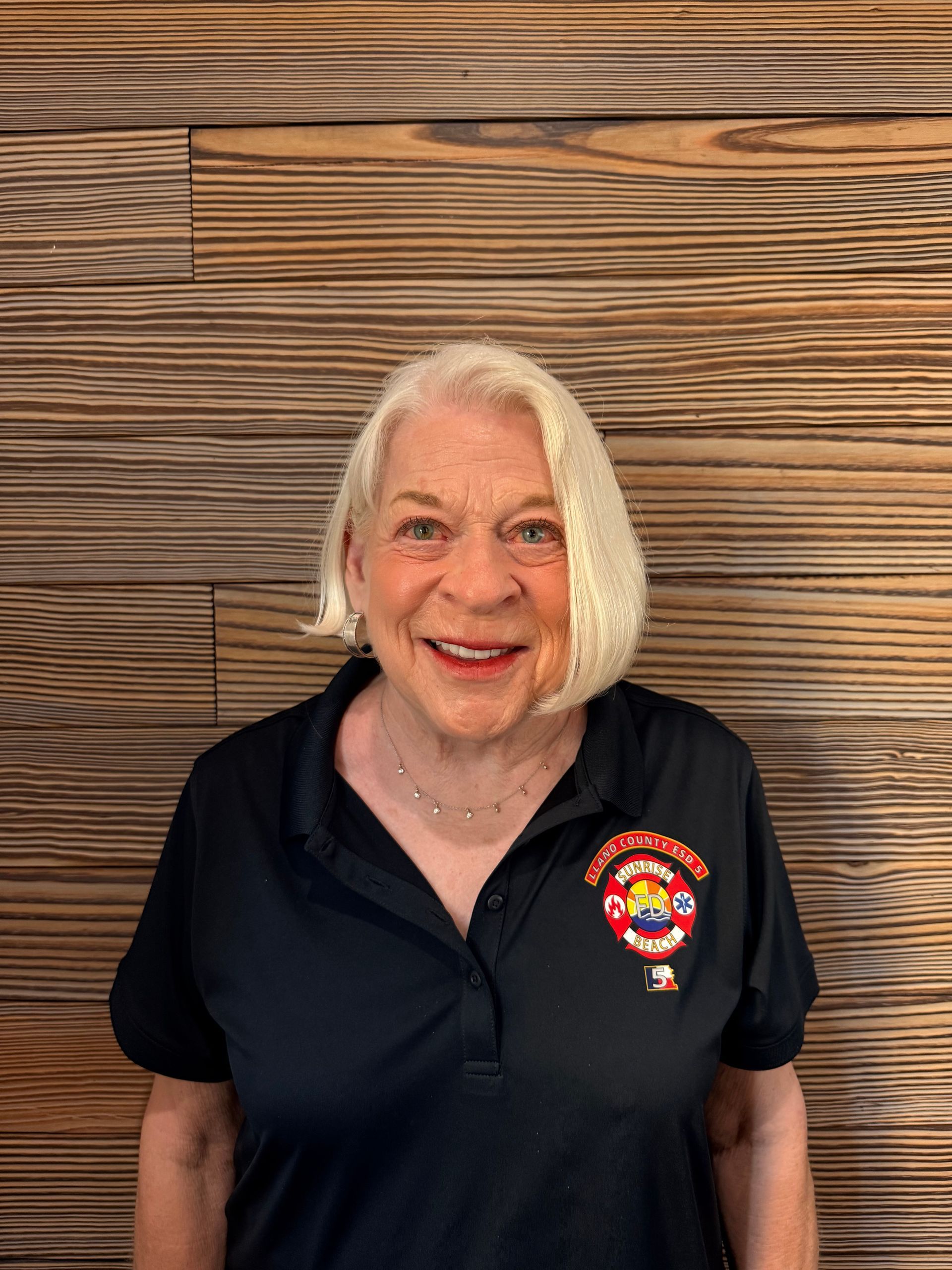 Smiling woman in a black polo with a colorful logo, standing before a wood-paneled wall.
