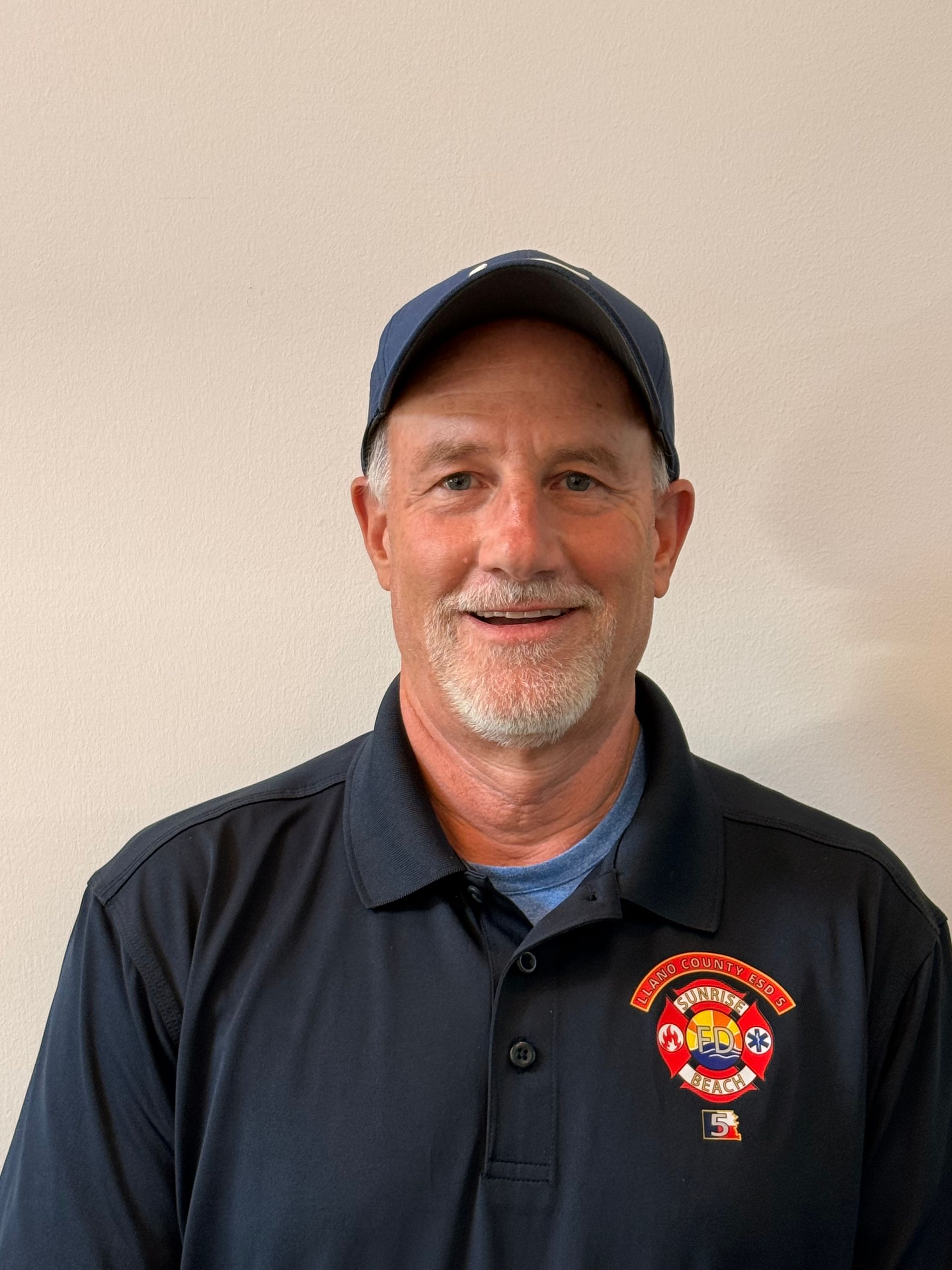Man in navy cap and polo shirt with embroidered logo, smiling against a plain beige wall