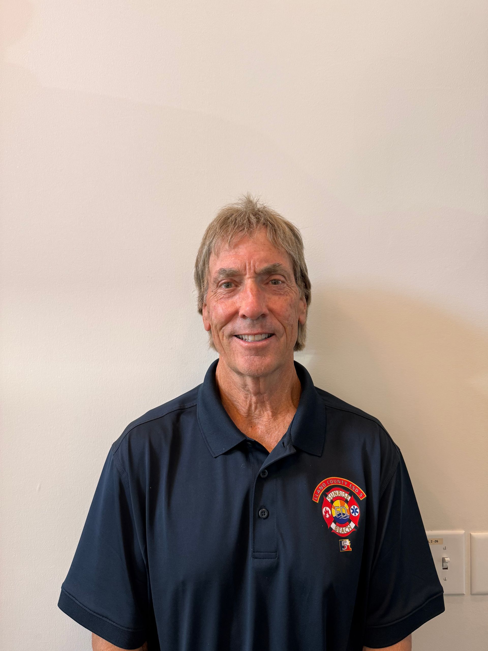 Smiling man in a navy polo shirt with a colorful emblem, standing against a plain wall.