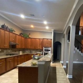 Large kitchen with wood cabinets, central island, tiled backsplash, and arched hallway opening into a stairway.