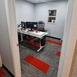 Small office with desk, monitors, chair, and gray carpet with red accents viewed through a doorway