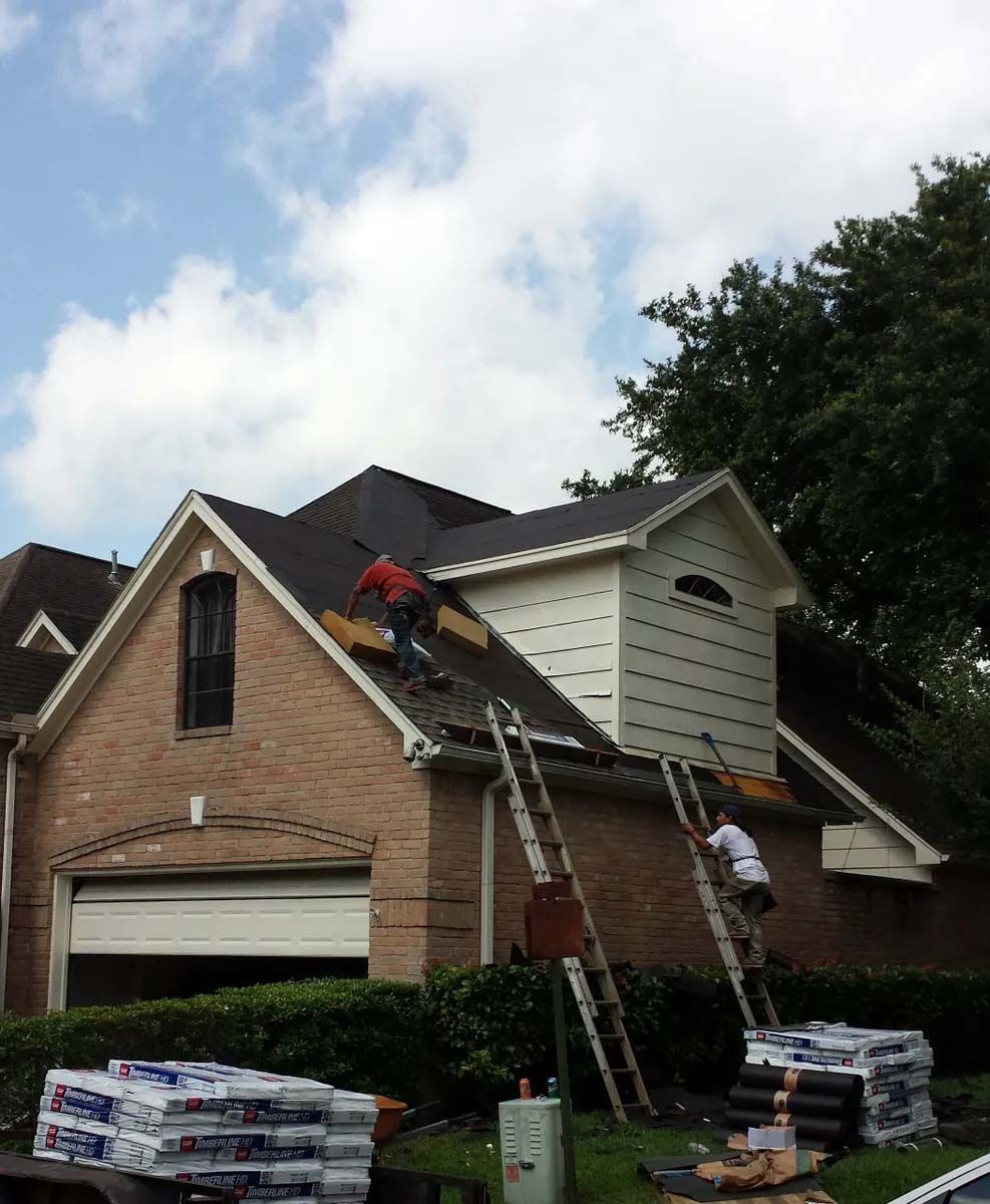 Two workers reroofing a brick house, with ladders and stacks of shingles in the front yard.