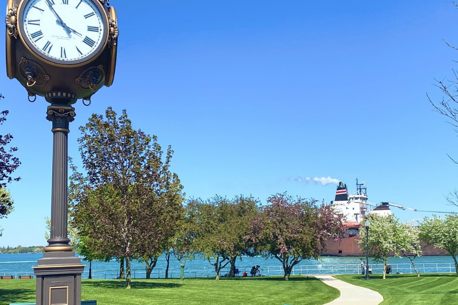 Clock in a park, trees with pink blossoms, people on a green lawn, and a building in the distance, under blue sky.
