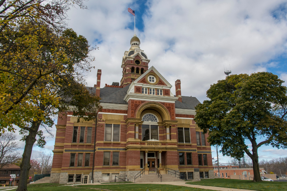 Brick courthouse with a central tower, flag, and trees on a cloudy day.