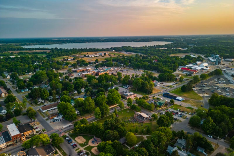 Aerial view of a town with trees, buildings, a lake, and a cloudy sky at sunset.