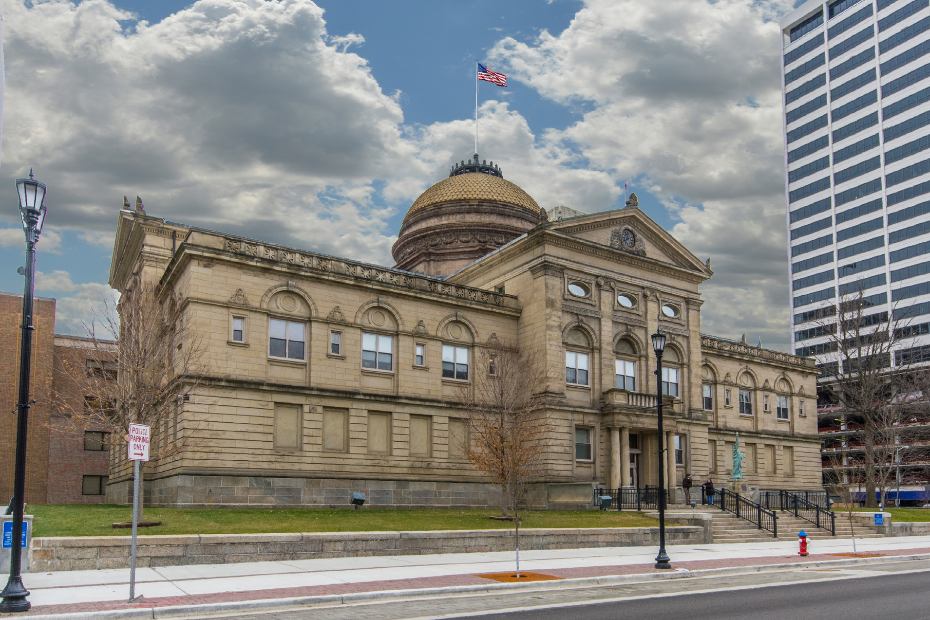 Beige courthouse with dome and flag under cloudy sky, next to a tall building.