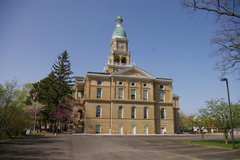 Beige courthouse with green-topped clock tower against blue sky, trees on sides, empty pavement in front.