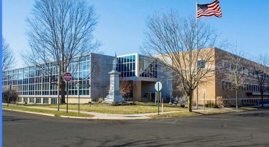 Two-story school building with glass and brick exterior, American flag, monument, trees, and street.