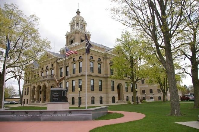 Beige courthouse with a clock tower and flags on a green lawn with a winding path.