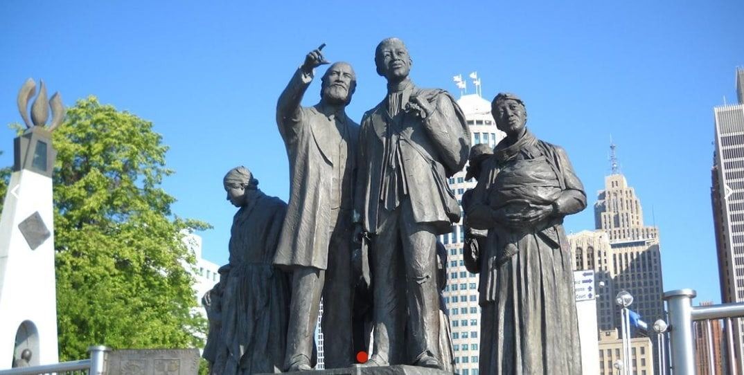Bronze sculpture group in Detroit; figures include pointing man and woman holding a basket; city skyline in background.