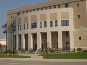Beige courthouse with curved facade, columns, windows, stairs, American flag, and grassy lawn.