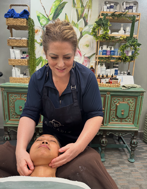 A spa professional in a dark apron gives a facial treatment to a client lying on a table in a treatment room.