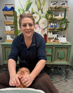 An aesthetician in a dark apron performs a facial massage on a client in a treatment room with floral wall art.