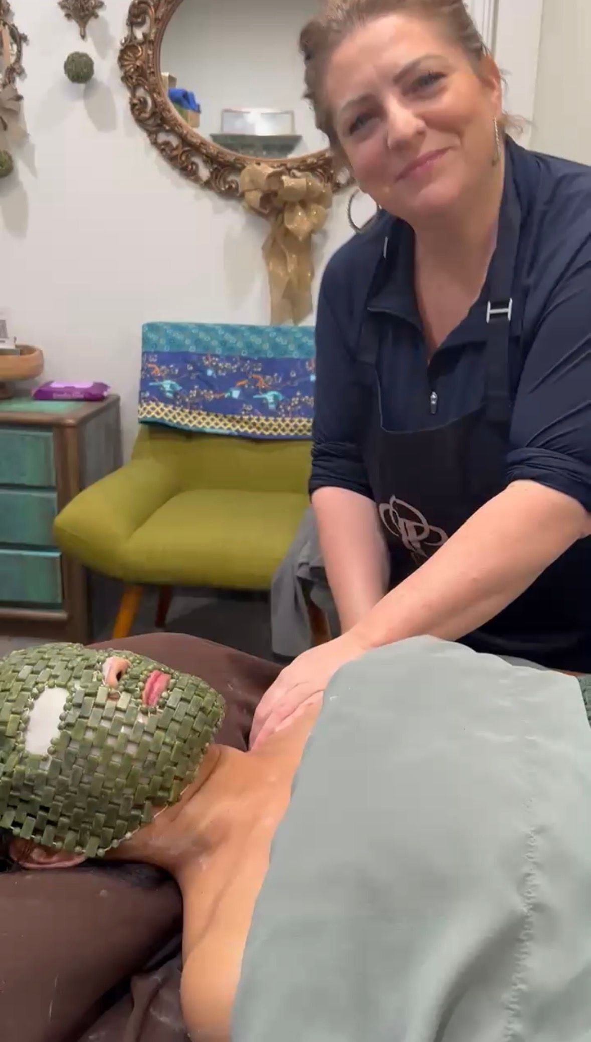 A professional performs a massage on a client wearing a textured green facial mask in a spa setting.