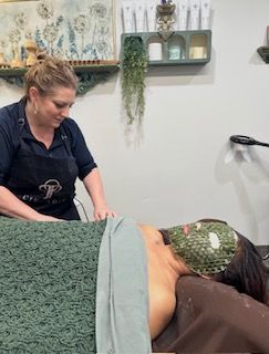 A professional aesthetician performs a facial treatment on a client wearing a textured green mask in a spa setting.