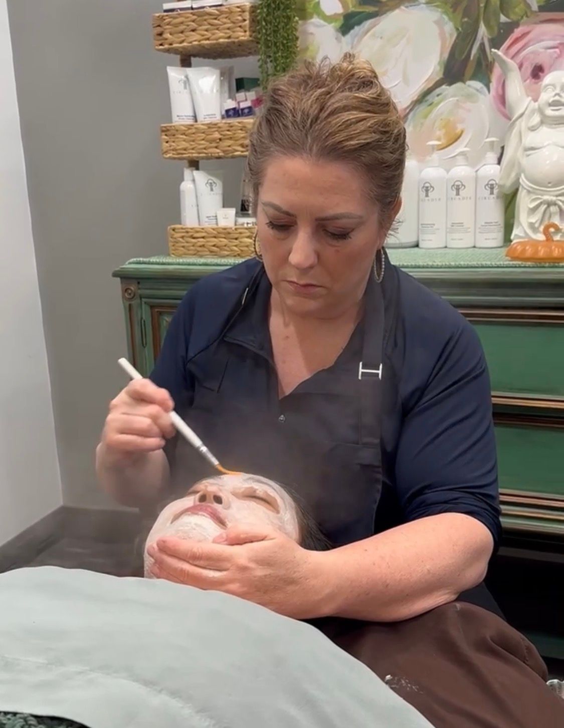 A professional applies a mask to a client's face with a brush while a steamer emits mist in a salon setting.