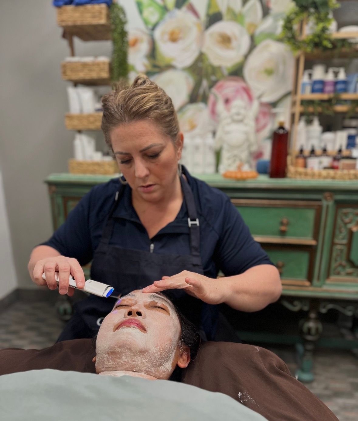 A professional performs a facial treatment on a client lying down, using a handheld device while applying product.