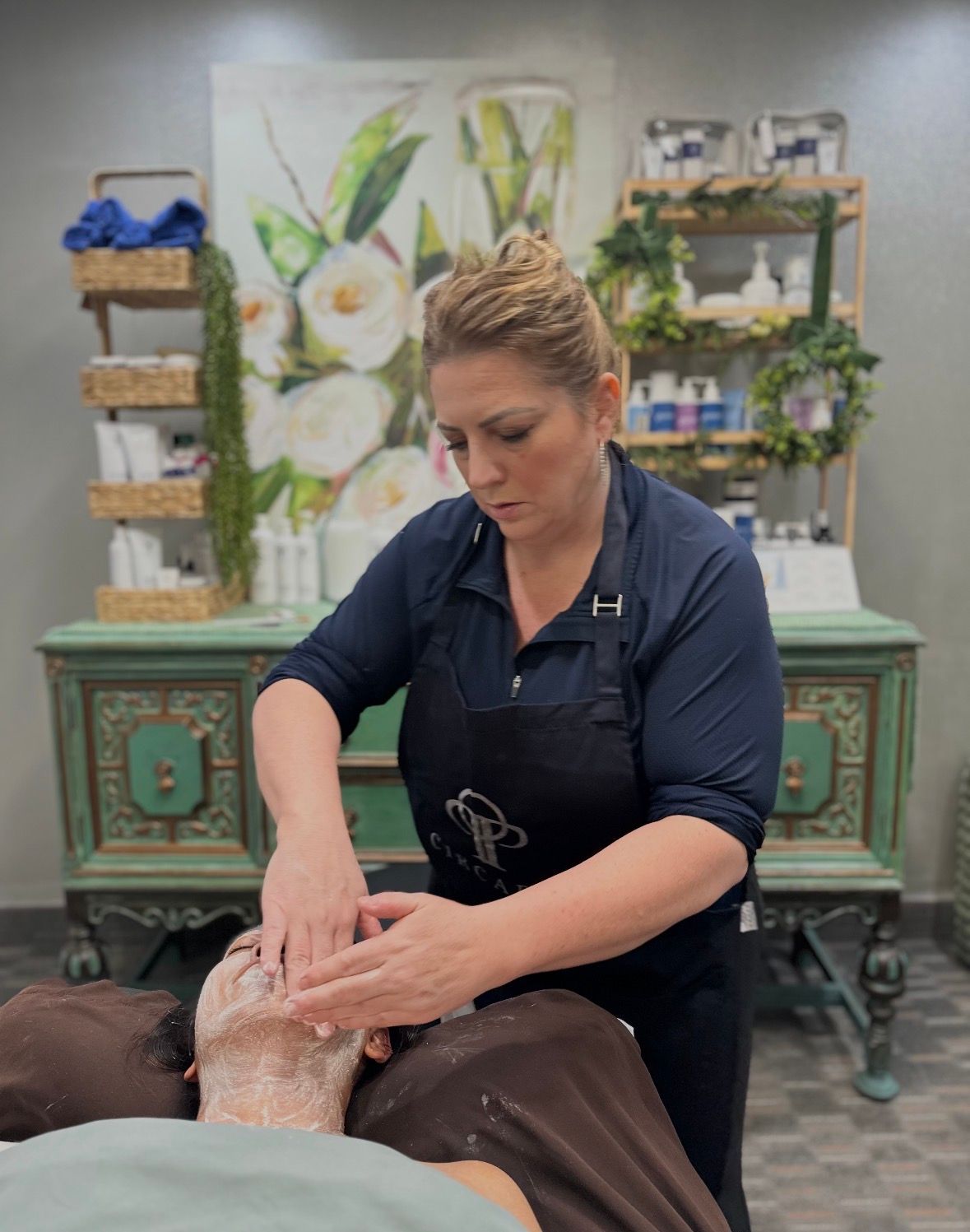 A professional providing a facial treatment in a spa setting with products on shelves in the background.