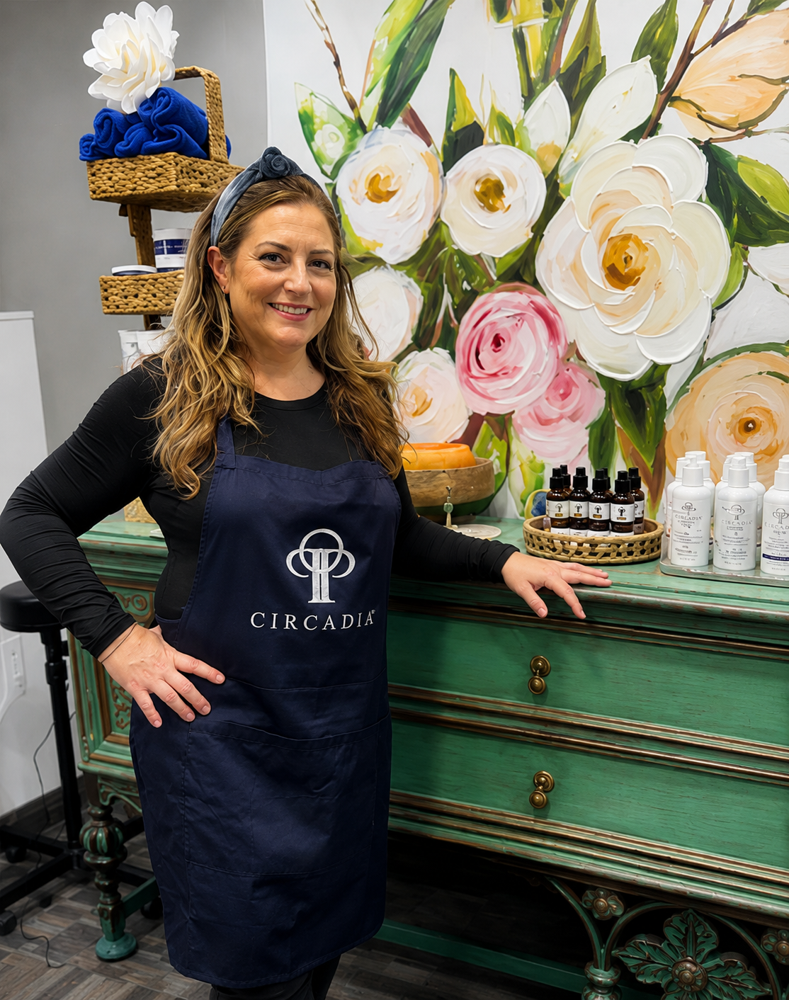 A smiling person wears a navy Circadia apron, standing in front of a floral mural and a green decorative cabinet.