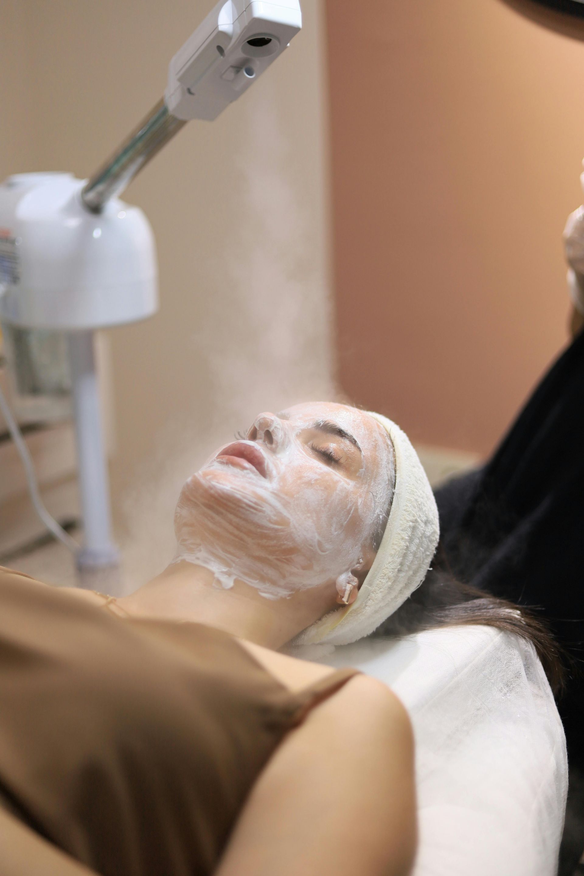 Woman receiving facial steam treatment at a spa, lying down with white mask on face.