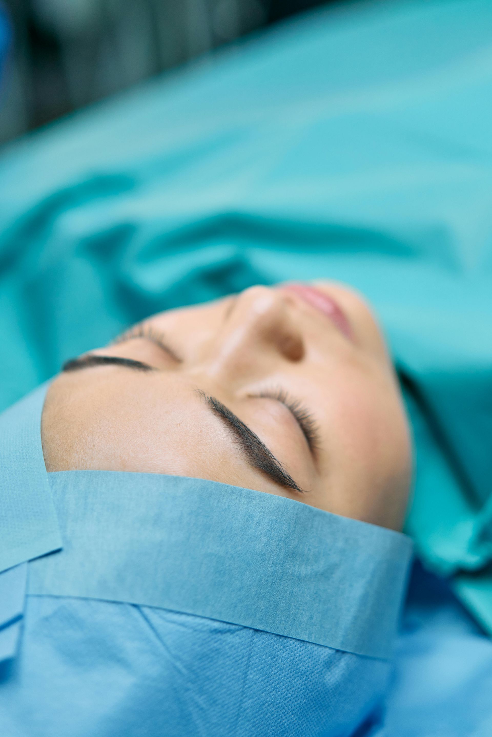 Person lying on operating table, eyes closed, wearing surgical cap and gown.