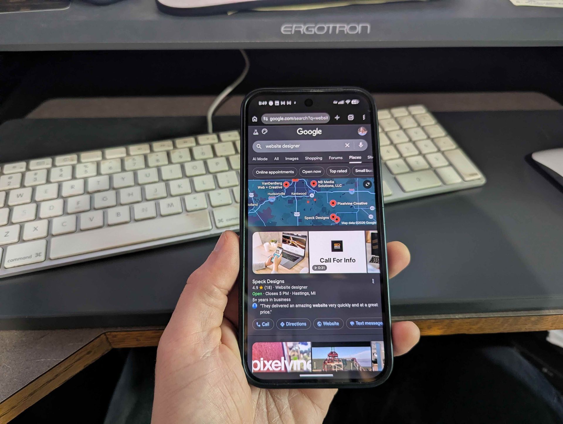 Hand holding a smartphone showing a dark-mode app screen in front of a white keyboard on a desk