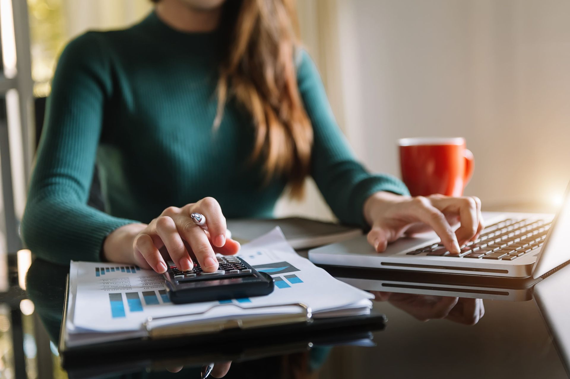 Woman using a calculator and laptop with financial reports on a desk.