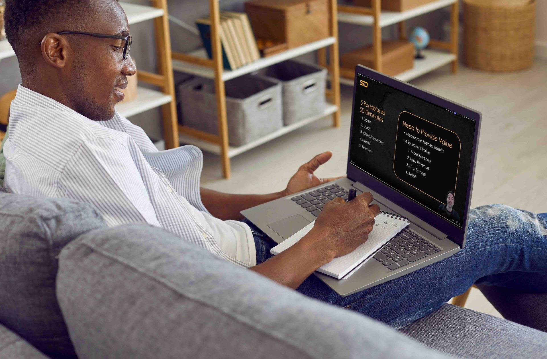 A man is sitting on a couch using a laptop computer, watching a video of Steven from Speck Designs explaining their services.
