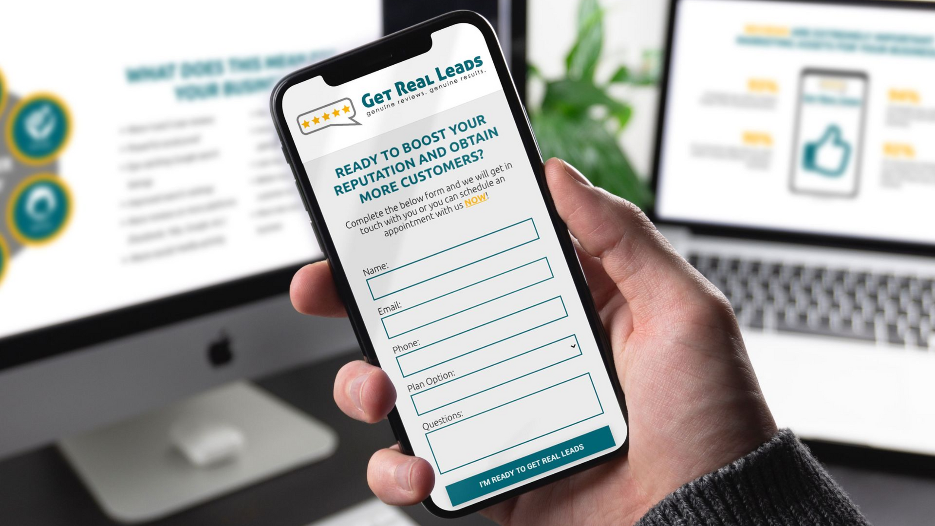 A hand holding a phone displaying a form for lead generation, with a laptop and monitor visible in the background.