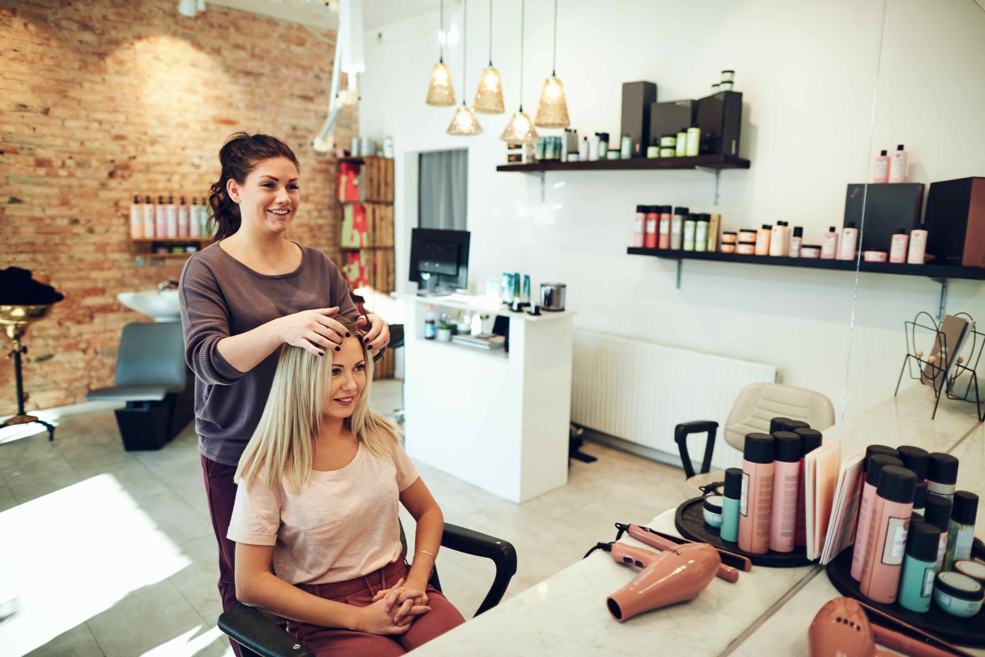 Hairdresser styling client's hair in a salon; blonde woman seated, brick wall background.