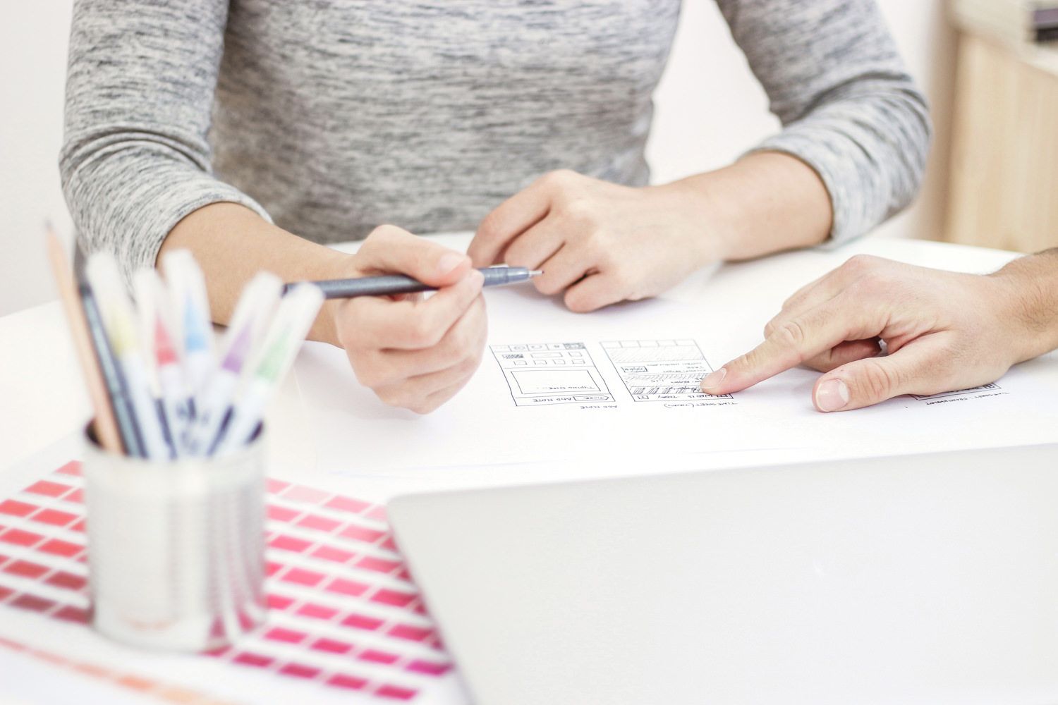 Two people at a table discussing sketches of a mobile app, pencils and a laptop nearby.