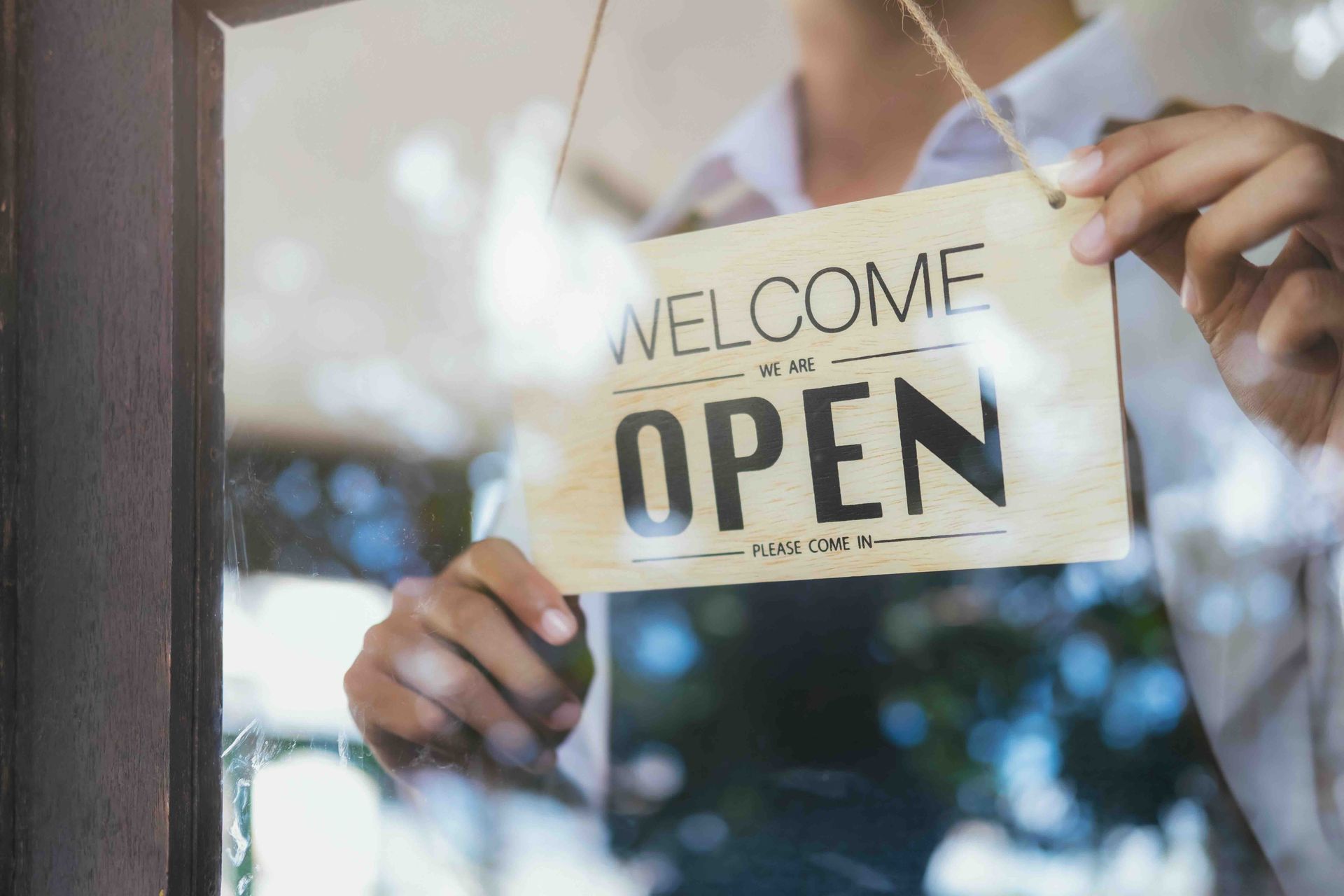 A person is holding a welcome we are open sign in front of a glass door.