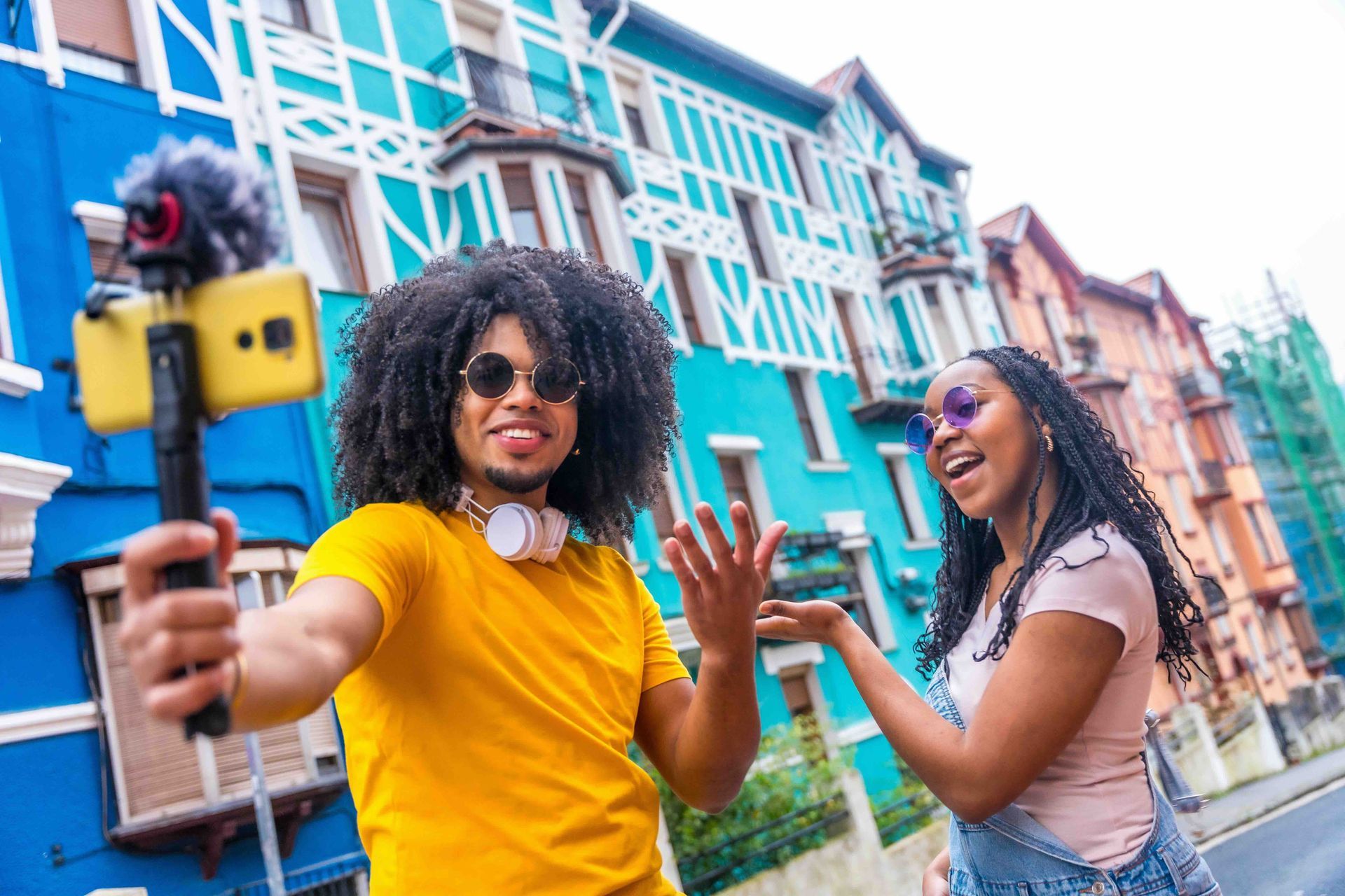 Two young people vlogging in front of colorful buildings. A man holds a phone with a mic, smiling. The woman gestures excitedly.