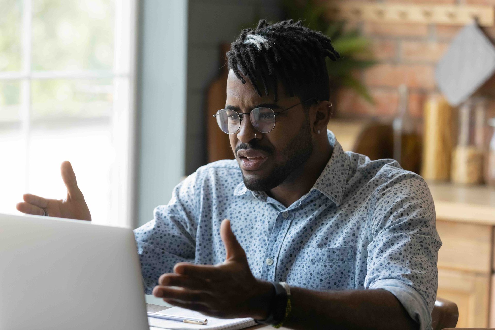 A man wearing glasses is sitting at a table using a laptop computer with a confused look on his face.